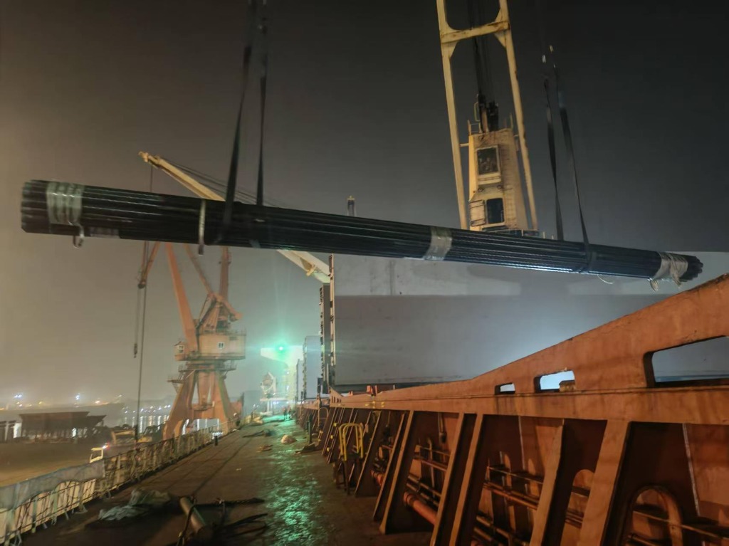 Night-time crane hoisting 12m API 5L 3LPE coated steel line pipes into vessel hold